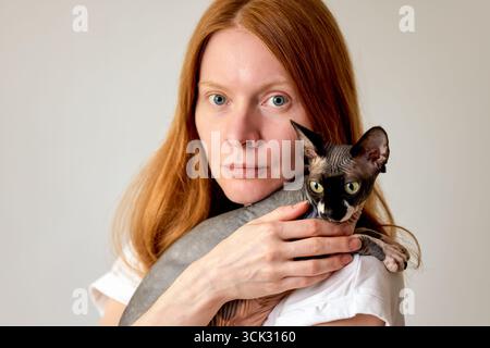 Eine rothaarige Frau umarmt ihre Sphynx-Katze und hebt Zuneigung und Gesellschaft hervor. Das Bild zeigt natürliches Licht und einen minimalistischen Hintergrund. Stockfoto