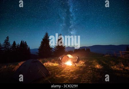 Silhouette von zwei Wanderern, die am Lagerfeuer unter Sternenhimmel ruhen, in der Nähe beleuchteter Zelte auf dem Campingplatz. Nächtliches Camping in den Bergen, Nachthimmel schaffen magische und friedliche Atmosphäre. Stockfoto