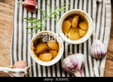Über dem Blick auf im Ofen geröstetes Knoblauchconfit in kleinen Ofentöpfen auf Küchentuch. Rosmarinzweig und Knoblauchhandschuhe zur Dekoration. Stockfoto