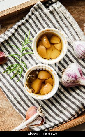 Über dem Blick auf im Ofen geröstetes Knoblauchconfit in kleinen Ofentöpfen auf Küchentuch. Rosmarinzweig und Knoblauchhandschuhe zur Dekoration. Stockfoto