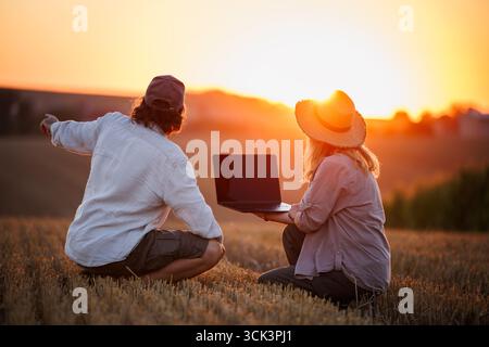Landwirte, die Laptop für intelligente Landwirtschaftsanalysen verwenden, während sie das ländliche Feld bei Sonnenuntergang beobachten. Konzept der modernen Landwirtschaft und Innovation in landwirtschaftlichen Betrieben Stockfoto