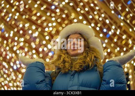 Wintermarkt-Szene mit jungen Frauen, die nach oben blicken, gebündelt in warmer Kleidung, umgeben von funkelnden weihnachtsbeleuchtung, die ein festliches, saisonales Ambiente schafft Stockfoto