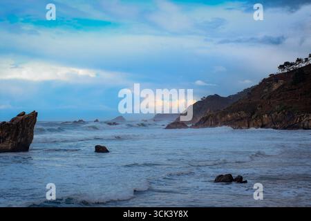 Eine wilde, dramatische Meereslandschaft mit einer felsigen Küste und mächtigen Wellen, die unter einem stürmischen, bewölkten Himmel gegen die Küste krachen Stockfoto