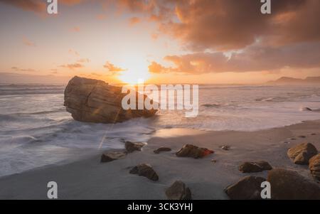 Der Blick auf die Sonnenstrahlen, die über einem monolithischen Felsen platzen, während schaumige Wellen am Sandstrand unter einem dramatischen Sonnenuntergangshimmel krachen, Kapstadt, Westkap, Südafrika. Stockfoto