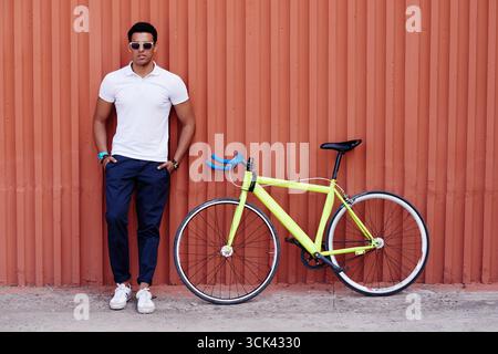 Junger erwachsener lateinischer Mann, der mit überkreuzten Armen neben gelbem Straßenfahrrad vor einer gewellten orangen Wand steht, eine Sonnenbrille trägt und selbstbewusst in die Kamera blickt Stockfoto