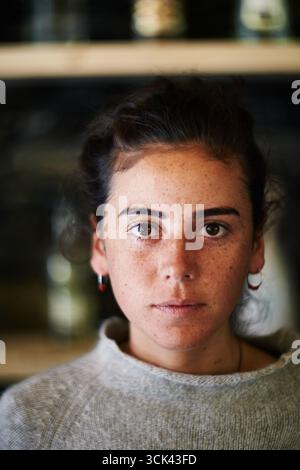 Porträt einer jungen Erwachsenen kaukasischen Frau, die direkt in die Kamera blickt, mit neutralem Ausdruck, dunklen Haaren zurückgezogen, sichtbaren Sommersprossen, einfachen Ohrringen und Pullover Stockfoto