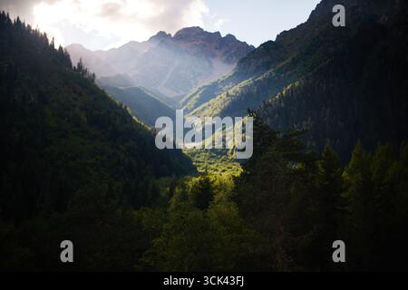 Berglandschaft mit sonnendurchflutetem Tal, umgeben von dichtem grünen Wald und hohen Gipfeln, mit dramatischem Sonnenlicht, das durch Wolken strömt und Hänge und Baumkronen beleuchtet Stockfoto