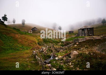 Ländliche Landschaft mit Holzhäusern, die über grasbewachsene Hügel mit Holzzäunen und einem kleinen Bach, der durch das Tal fließt, verstreut sind, Nebel bedeckt entfernte Bäume und Hänge Stockfoto