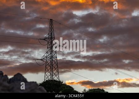 Hochspannungsmasten gegen den Himmel während Sonnenuntergang, Stromleitung, Sonnenuntergang mit bewölktem orangefarbenen Himmel Stockfoto