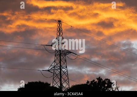 Hochspannungsmasten gegen den Himmel während Sonnenuntergang, Stromleitung, Sonnenuntergang mit bewölktem orangefarbenen Himmel Stockfoto