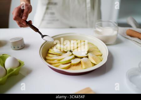 Unbekannte Frau streut Zucker über geschnittene Äpfel in der Backform, bereitet Dessert in der Küche, Eier und Glasschale mit Zucker sichtbar auf der Arbeitsplatte Stockfoto