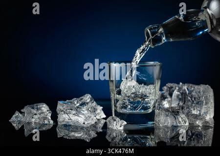Kristallklarer Wodka oder Gin wird über Eis in ein Glas gegossen. Wodka und Eisstücke auf schwarzem reflektierendem Hintergrund. Kopierbereich. Stockfoto