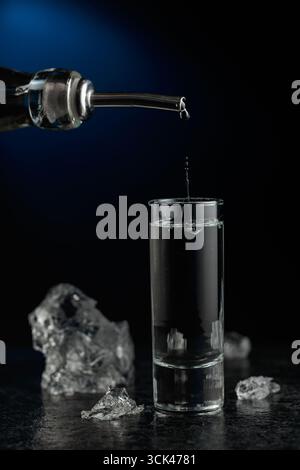 Kristallklarer Wodka oder Gin wird in ein Glas gegossen. Transparentes alkoholisches Getränk mit Eisstücken auf schwarzem Hintergrund. Stockfoto
