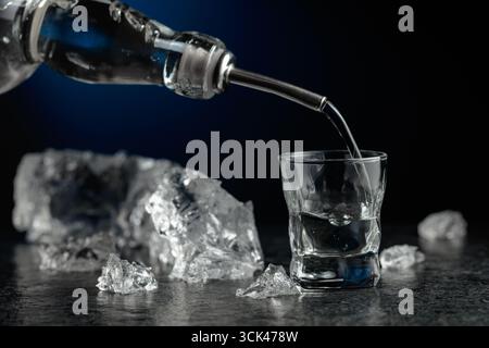 Kristallklarer Wodka oder Gin wird in ein Glas gegossen. Transparentes alkoholisches Getränk mit Eisstücken auf schwarzem Hintergrund. Stockfoto