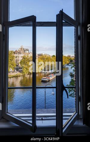 Blick aus einem Fenster auf ein Frachtschiff auf der Singelgracht Kanal, Amsterdam, Niederlande. Blick aus einem Fenster auf ein Frachtschiff auf der Singelgracht, Stockfoto