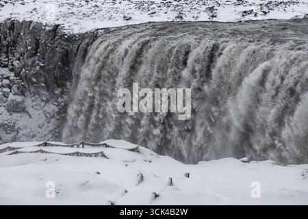 Blick auf einen mächtigen Wasserfall, der über eine felsige Klippe in eine schneebedeckte Landschaft stürzt, und der eisige Sprühnebel erzeugt einen nebeligen Schleier in Island, Island. Stockfoto