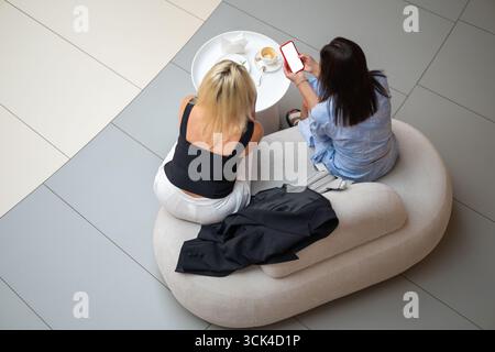 Zwei junge Frauen sitzen auf einer gemütlichen Couch an einem Food Court-Tisch im Einkaufszentrum, plaudern bei Kaffee und teilen Informationen auf einem Smartphone von oben nach unten Stockfoto