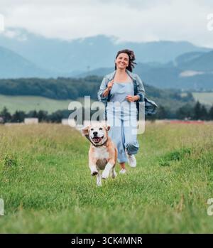 Glückliche lächelnde Beagle Hund laufen Porträt Zunge aus mit weiblichen Besitzer joggen entlang Bergwiesen Gras Pfad Outdoor Walking Natur Haustiere gesund ac Stockfoto