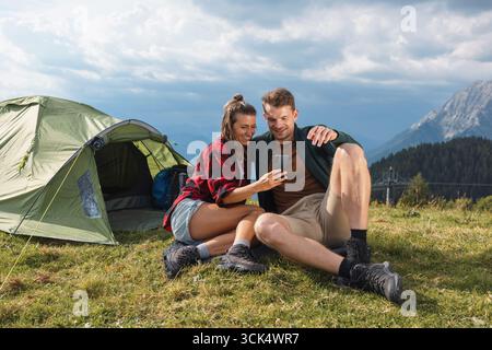 Romantisches Camping-Paar, das mit einem Smartphone Momente im Freien festnimmt, sitzt neben einem Zelt in einer malerischen Berglandschaft. Stockfoto