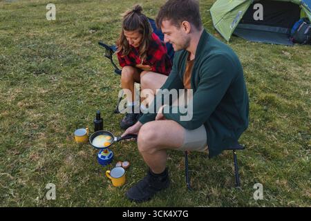 Zwei Personen kochen das Frühstück am Lagerfeuer in einem grasbewachsenen Bereich mit einem Zelt im Hintergrund an einem sonnigen Morgen. Stockfoto