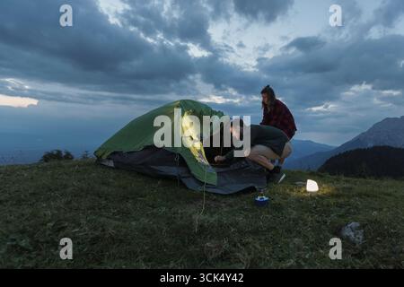 Ein Paar baut ein Zelt in der bergigen Landschaft auf, während die Dämmerung einbricht, beleuchtet von sanftem Licht einer Laterne, umgeben von der Schönheit der Natur. Stockfoto