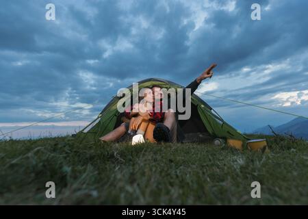 Zwei Leute sitzen zusammen in einem Zelt auf einem grasbewachsenen Hügel und zeigen in der Dämmerung auf die Wolken. Die Atmosphäre ist entspannt und intim. Stockfoto