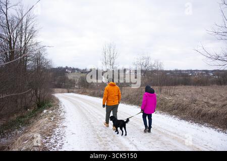 Ein Paar geht Hand in Hand, ein verspielter Hund an der Seite, entlang eines verschneiten Pfades in der Landschaft, umgeben von braunen Feldern und fernen Hügeln unter einem cl Stockfoto