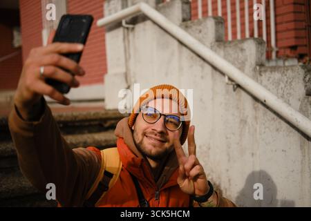 Ein lächelnder Mann in einer Brille, der Selfie macht, mit den Fingern draußen ein Friedenszeichen zeigt. Reiseblogger-Konzept. Stockfoto