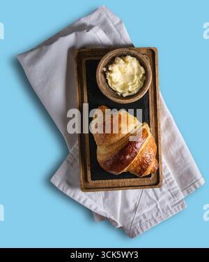 Ein frisch gebackenes Croissant, serviert auf einem Holztablett mit einer Schüssel Butter, ideal zum Frühstück oder für einen Snack. Stockfoto