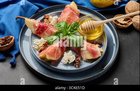 Birnenkeile in Schinkenschinken gewickelt und mit Sesamsamen bestreut, serviert mit einer kleinen Schüssel Honig und einem hölzernen Honiglöffel auf einer blauen Keramikplatte. Stockfoto
