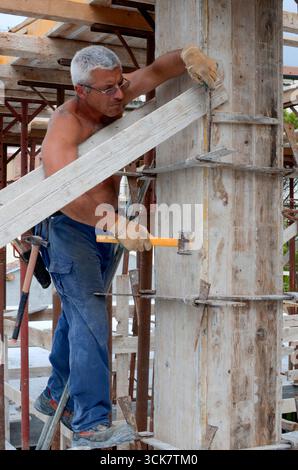 Ein fokussierter Baumeister arbeitet mit einem Hammer an einem Holzrahmen, der Bauarbeiten, Handarbeit und Hingabe in der Branche repräsentiert. Die Aufnahme hebt hervor Stockfoto