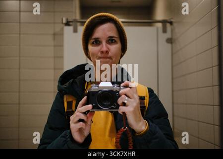 Frau in gelber Mütze und Jacke, die ein Spiegel-Selfie mit einer Retro-Kamera in öffentlichen Toiletten macht. Stockfoto