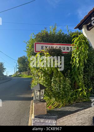Ruhige Straße in Exincourt, Frankreich, mit einer geschwungenen Straße, Steinmauer, Stadtschild, charmanten Häusern, kleinen Gärten, üppiges Grün und sonniger blauer Himmel Stockfoto