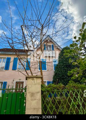 Bezauberndes französisches Haus mit hellblauen Fensterläden, verziertem Balkon und Gartenbäumen unter einem teilweise bewölkten Frühlingshimmel. Stockfoto