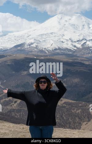 Frau in schwarzem Pullover und Hut genießt einen Panoramablick auf den schneebedeckten Mount Elbrus unter einem dramatischen Himmel Stockfoto