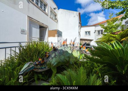 Eine farbenfrohe Dinosaurier-Skulptur steht inmitten üppiger grüner Farne in einem sonnigen Innenhof, umgeben von weißen Gebäuden unter einem hellblauen Himmel in Lourinhã Stockfoto