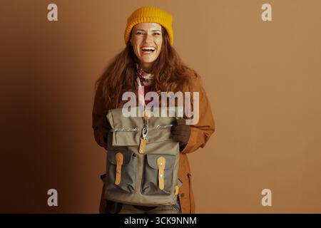 Eine fröhliche Frau mit langen braunen Haaren, mit gelber Mütze, braunem Mantel und Schal, strahlt in die Kamera, während sie einen grünen Rucksack hält Stockfoto