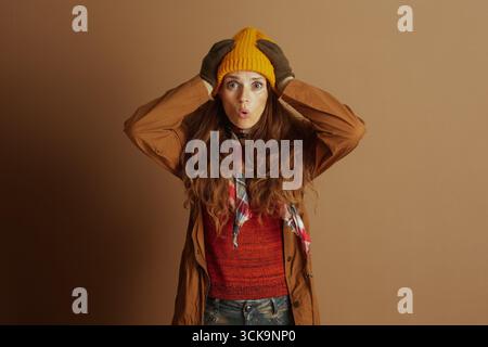 Eine Frau in kuscheliger Herbst-Winter-Kleidung, einschließlich Trenchcoat und Mütze, schaut den Betrachter mit einem breiten, überraschten Ausdruck, der den Betrachter direkt ansieht Stockfoto
