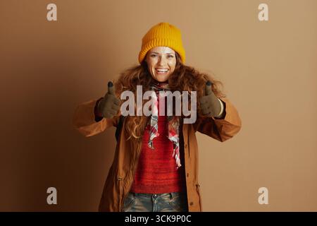 Das fröhliche Porträt einer Frau in gelber Mütze, rotem Pullover und gemustertem Schal sorgt für doppelte Daumen nach oben. Ihre warme Kleidung und ihr breites Lächeln deuten auf Readines hin Stockfoto