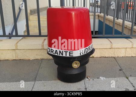 Roter Hydrant mit schwarzem Sockel, auf einem Bürgersteig in der Nähe eines Gebäudes, der die städtische Infrastruktur und Sicherheitseinrichtungen in einer städtischen Umgebung darstellt Stockfoto