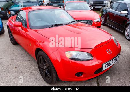 Roter Mazda MX-5 Sportwagen parkt an einem sonnigen Tag auf einem Parkplatz mit verschiedenen anderen Fahrzeugen im Hintergrund Stockfoto