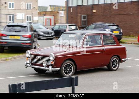 Ein braunes Oldtimer austin A40 fährt auf einer Straße mit modernen Fahrzeugen, die im Hintergrund geparkt sind und eine Mischung aus klassischem und zeitgenössischem Stil bietet Stockfoto