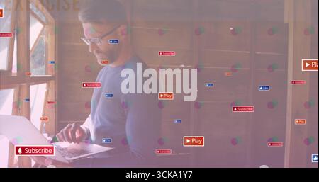 Mann tippt auf silbernem Laptop im Büro durch hölzernen Fensterrahmen, digitale Überlagerungssymbole, Kopierraum Stockfoto