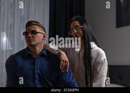 Ein Mann in einem blauen Hemd und eine Frau in einem weißen Hemd stehen dicht beieinander und tragen eine Brille. Sie befinden sich in einem gut beleuchteten Innenbereich und strahlen eine schicke Atmosphäre aus, während sie für die Kamera posieren. Stockfoto