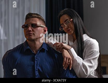 Ein Mann mit blauem Hemd und eine Frau mit weißem Hemd sitzen dicht beieinander. Sie setzen sich in einer stilvollen Innenumgebung mit sanfter Beleuchtung selbstbewusst auf. Stockfoto