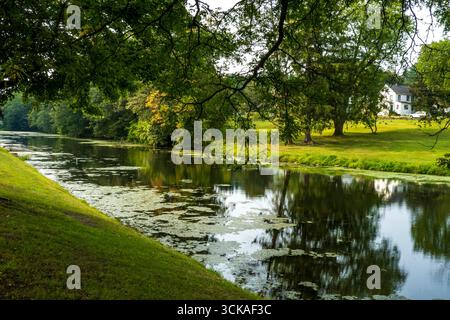 Monroe, NY - USA - 6. September 2025 : ruhige Teichszene in Monroe, NY, mit üppigen grünen Ufern, reflektierendem Wasser und einem entfernten weißen Haus unter einem Clo Stockfoto