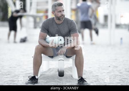 Sportmann, der draußen auf einer Bank ruht. Reifes Fitnesssportmodell, das nach dem Training auf einer Bank sitzt. Sportmann der 40er Jahre, der nach dem Sport eine Pause macht. Passen Stockfoto