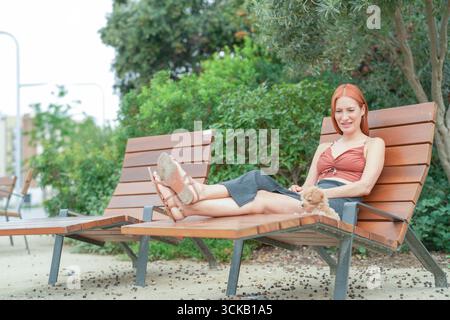 Eine Frau sitzt auf einer Bank mit erhobenen Füßen und ein Hund neben ihr. Die Szene ist friedlich und entspannend, die Frau genießt die Natur und die Stockfoto