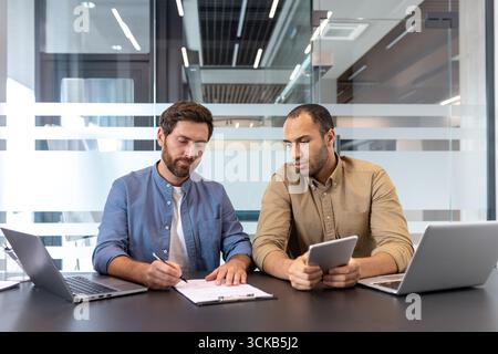 Zwei Geschäftsleute arbeiten an einem Projekt zusammen, einer schreibt ein Dokument, der andere hält ein digitales Tablet, das Teamwork und Technologie in einem modernen Büro verkörpert Stockfoto