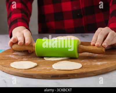 Ein Kind rollt den Teig mit einem Drehstift auf einem Holzbrett aus, Nahaufnahme Stockfoto